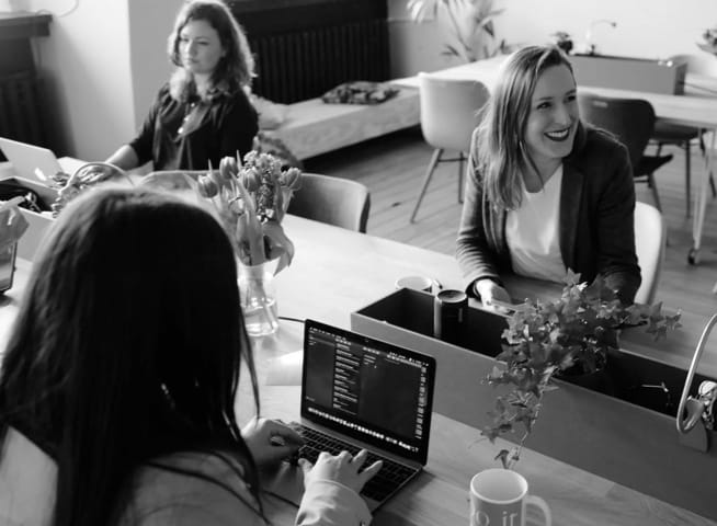 Three women sitting at one big desk working behind computers
